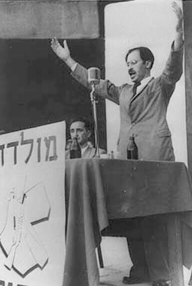 מנחם בגין נואם לאחר יציאתו מהמחתרת, ב-14 באוגוסט 1948, בתל אביב. יושב לידו חיים לנדאו. מלפניו הכתובת "מולדת וחרות" וסמל אצ"ל. מתוך ויקיפדיה