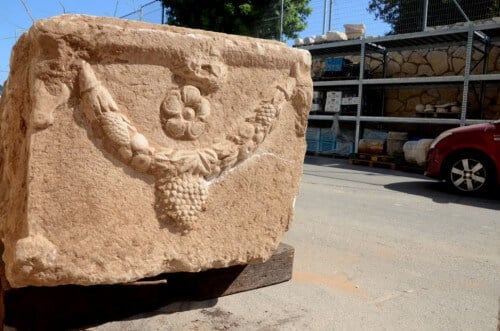 A wreath emerging from the sides of the high edges of the sarcophagus, decorated with bulls' heads, the wreath is made of acanthus leaves and pine cones and fruits are combined in it. Its center carries a bunch of grapes and in its field a rose-like decoration is displayed.