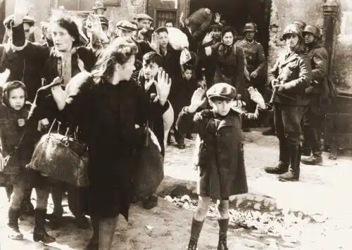 A boy raises his hands during the removal of Jews from the bunkers during the suppression of the uprising in Warsaw, 1943. The boy has been identified as Artur Domb Semyontek, Israel Rundel, Zvi Nussbaum or Levi Zelinorger. German Federal Archives / Public domain