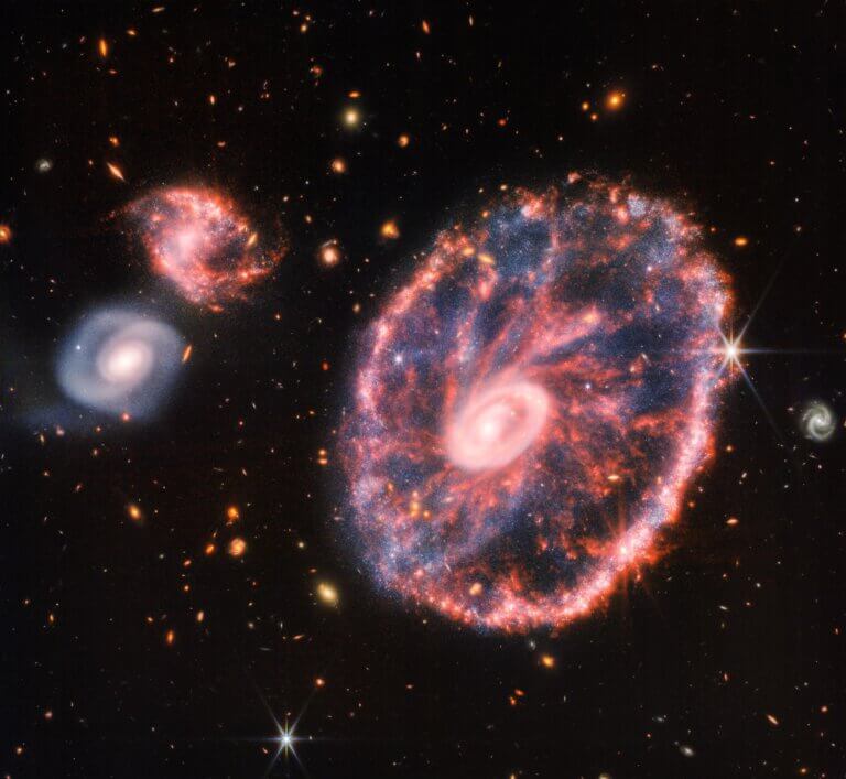 A large spotted pink wheel-like galaxy with a small inner ellipse, with a hazy blue in the middle to the right, with two smaller spiral galaxies of about the same size to the left on a black background. This image of the Wheel Galaxy and its companion galaxies is a composite from the Near Infrared Camera and Webb's Mid-Infrared Instrument, which reveals details that are difficult to see from the separate images alone. Credit: NASA, ESA, CSA, STScI