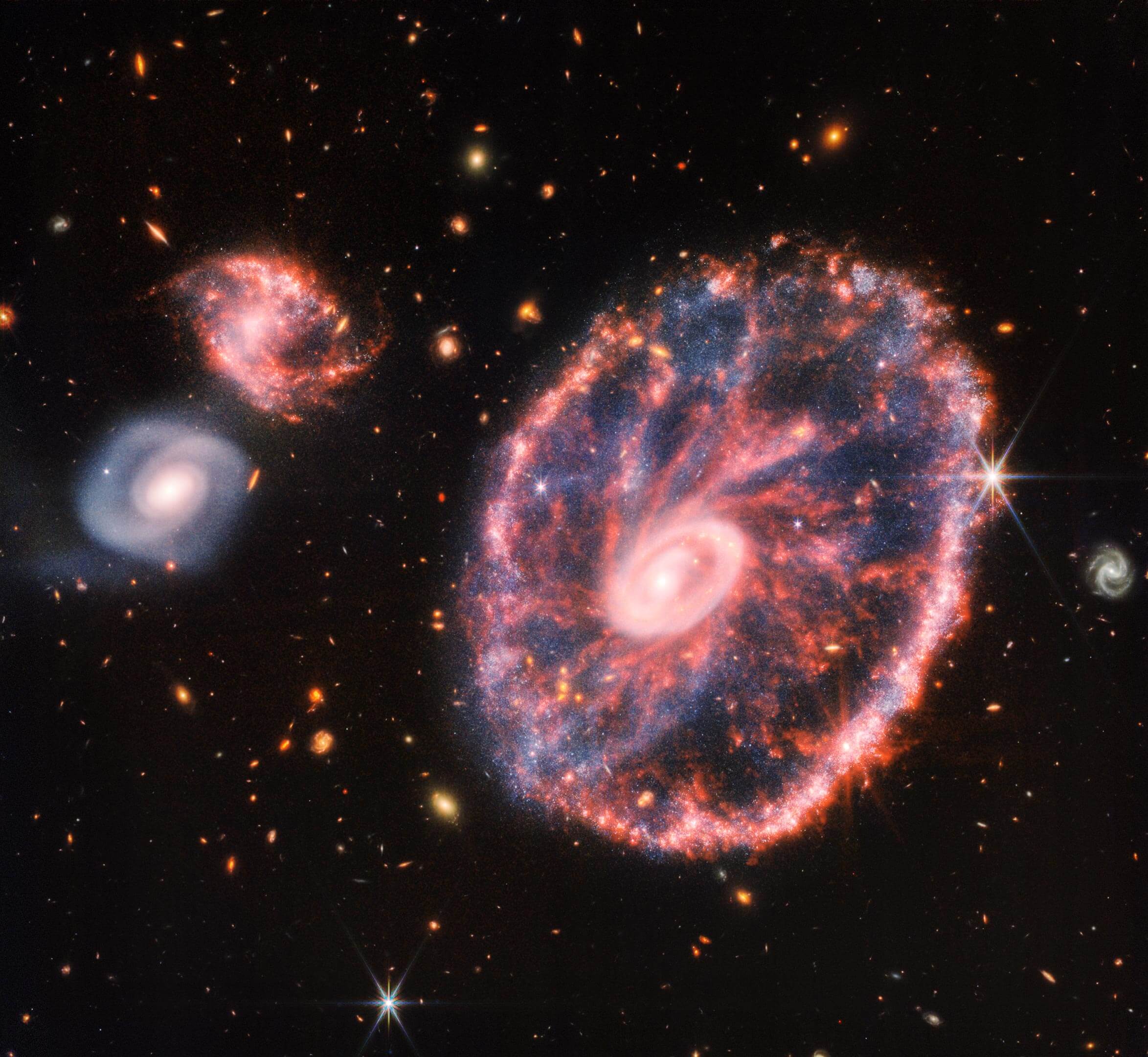 גלקסיה ורודה גדולה מנוקדת הדומה לגלגל עם אליפסה פנימית קטנה, עם כחול מעורפל בתווך מימין, עם שתי גלקסיות ספירליות קטנות יותר בערך באותו גודל משמאל על רקע שחור. תמונה זאת של גלקסיית הגלגל והגלקסיות הנלוות שלה היא הרכבה ממצלמת האינפרה אדום הקרוב וממכשיר האינפרה אדום הבינוני של ווב, שחושפת פרטים שקשה לראות מהתמונות הנפרדות לבדן. קרדיט: NASA, ESA, CSA, STScI