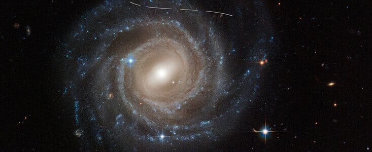 صورة تلسكوب هابل الفضائي للمجرة الحلزونية المحظورة UGC 12158. المجرة المهيبة لها شكل دولاب الهواء يتكون من نجوم زرقاء لامعة تحيط بتركيز أصفر-أبيض من النجوم المركزية. يحتوي هذا التركيز على خط قطري من النجوم، يسمى الشريط. تميل المجرة إلى الداخل بالنسبة لنا من الأرض. الخط الأبيض على شكل حرف S قليلاً عبر الجزء العلوي هو صورة هابل لكويكب يحوم عبر مجال الرؤية. يبدو متقطعًا لأن الصورة مركبة من عدة تعريضات للكويكب الذي يطير مثل سيارة السباق. المصدر: NASA، ESA، PG Martín (جامعة مدريد المستقلة)، J. DePasquale (STScI). المصدر: A. Filippenko (جامعة كاليفورنيا، بيركلي).