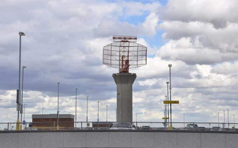 A tall radar tower, cloudy sky, light poles and buildings in an industrial complex.