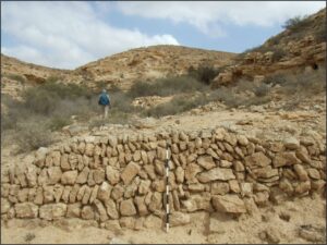 An explorer in a rocky desert terrain with an ancient, imposing stone wall in natural tones and a bright sky.