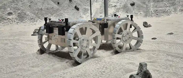 רכב רובוטי ניסויי בעל ארבעה גלגלים מעוקלים נע על משטח חול.