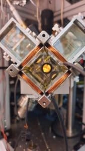 Caption: The 2D MOT system that delivers cold atoms to the experimental chamber. In the center is a glass chamber held in a vacuum between coils that create magnetic fields. The atoms pass into the experimental chamber through the tiny hole visible in the center of the yellow reflective surface. Credit: Or Dubkovsky.